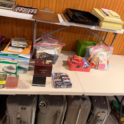 Sale Photo Thumbnail #102: Assorted office and household supplies arranged on folding tables, including legal pads, envelopes, and calculators, with several rolling suitcases stowed underneath.