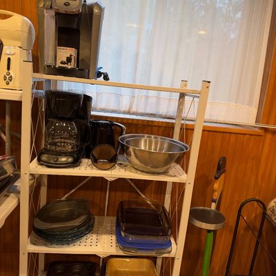 Sale Photo Thumbnail #108: White metal four-tier shelving unit loaded with various kitchen items, including a Keurig single-serve coffee maker, a Mr. Coffee drip coffee pot, stainless steel mixing bowls, glass baking dishes, and muffin tins.
