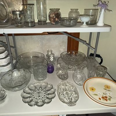 Sale Photo Thumbnail #76: Collection of vintage glass and ceramic kitchenware displayed on folding tables, including clear cut-glass bowls, an deviled egg platter, glassware, and stoneware floral plates.