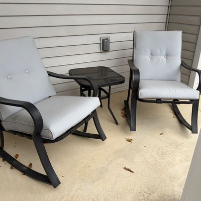 Sale Photo Thumbnail #93: Three-piece outdoor patio set including two black metal motion rocking chairs with light grey cushions and a matching glass-top side table.