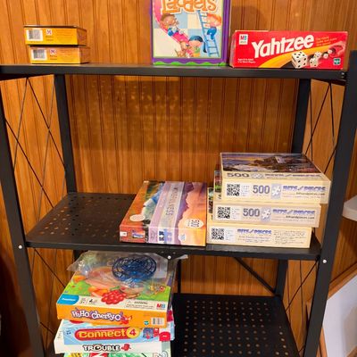 Sale Photo Thumbnail #112: Black metal four-tier shelving unit displaying a collection of classic board games and jigsaw puzzles, including Chutes and Ladders, Yahtzee, Connect 4, and Bits & Pieces puzzles.