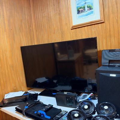 Sale Photo Thumbnail #99: Collection of home electronics on a folding table, including a large flat-screen TV, DVD/Blu-ray players, remote controls, portable radio, speaker, and small clip-on fans.