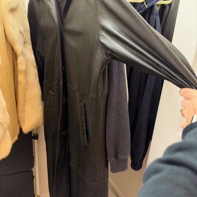 Sale Photo Thumbnail #55: Vintage long black leather trench coat hanging in a closet alongside other outerwear. The coat features side pockets, a collared neck, and a full-length silhouette.