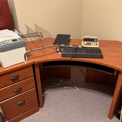 Sale Photo Thumbnail #47: Large warm wood-tone corner computer desk with a matching three-drawer pedestal cabinet, holding a printer, keyboard, and office accessories.