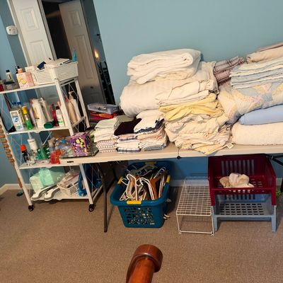 Sale Photo Thumbnail #29: Large assortment of household linens, blankets, and towels displayed on folding tables, accompanied by a shelving unit with toiletries and under-table storage baskets.