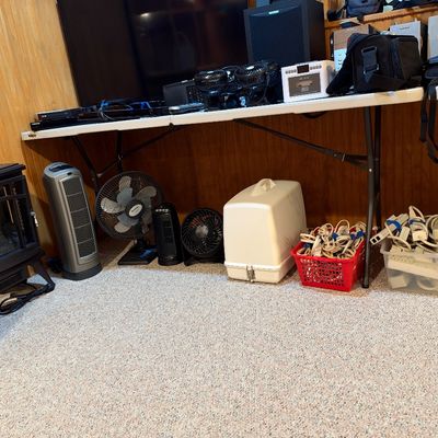 Sale Photo Thumbnail #97: Collection of household electronics including portable fans, space heaters, power strips, and a vintage sewing machine case displayed on a folding table and floor.