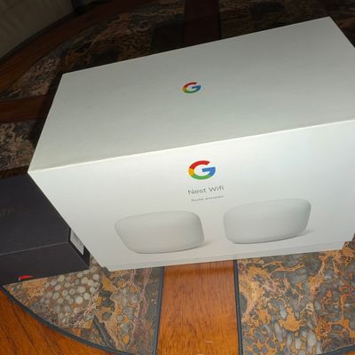 Sale Photo Thumbnail #69: Google Nest Wifi router and point system in original packaging, set alongside a smaller black electronics box on a patterned tabletop.