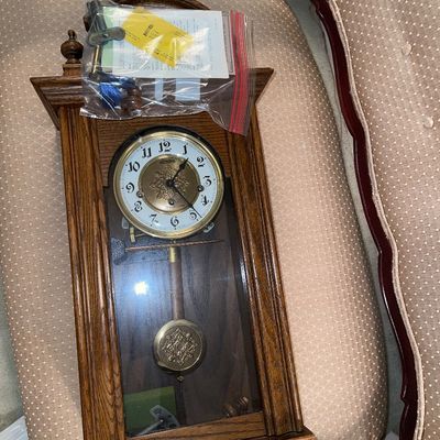 Vintage Linden wooden wall regulator clock featuring a brass pendulum and ornate face details, resting on a padded bench.