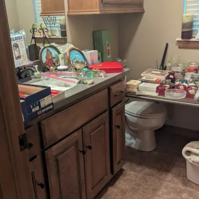 Bathroom counter and temporary shelving unit filled with various household items, glassware, small decor, and vanity accessories.