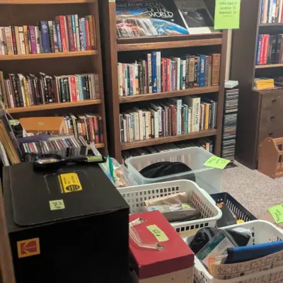 Collection of wooden bookcases filled with hardback and paperback books, atlases, and storage bins containing miscellaneous household items.