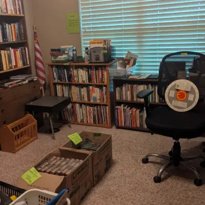 Home office and library layout featuring multiple wooden bookcases filled with books, a black mesh office chair, and boxes of media...