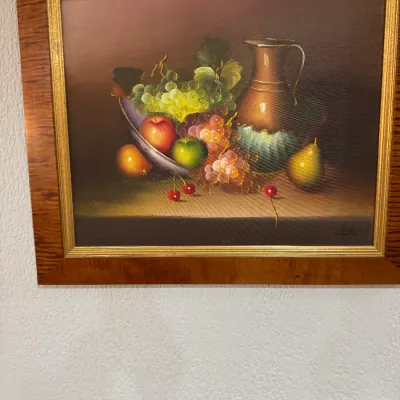 Framed still life oil painting depicting a bowl of fruit, grapes, cherries, and a pitcher, housed in a tiger maple wood frame with a gilt...
