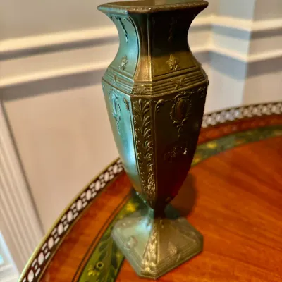 Antique-style bronze or brass urn vase featuring intricate relief details including fleur-de-lis motifs, laurels, and a square pedestal...