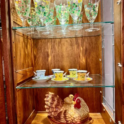 Display cabinet filled with hand-painted butterfly glassware, butterfly espresso cups with saucers, and a ceramic hen with chick covered...