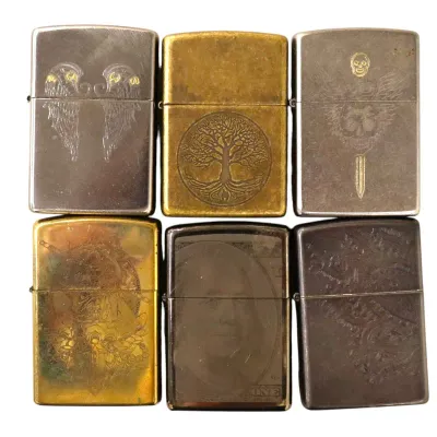 Sale Photo Thumbnail #160: Collection of six engraved metal flip-top lighters featuring designs including a tree of life, winged skulls, and a Benjamin Franklin hundred-dollar bill graphic.