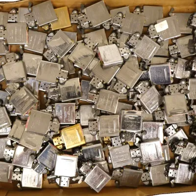 Sale Photo Thumbnail #197: Cardboard box filled with dozens of vintage metal cigarette lighter inserts, mostly Zippo and similar brands in silver and gold tones.