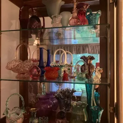 Sale Photo Thumbnail #7: Collection of vintage art glass pieces displayed in a wooden curio cabinet, including Fenton glass baskets, ruffled vases, and perfume bottles in various colors.
