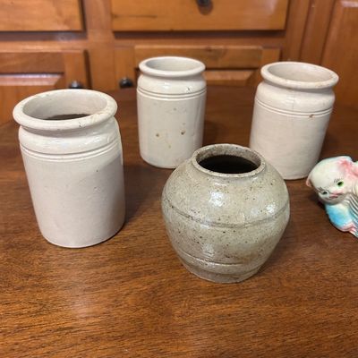 Sale Photo Thumbnail #22: Collection of four antique ceramic jars, including three matching off-white stoneware crock jars and one round speckled clay pot, displayed alongside a vintage porcelain animal figurine.