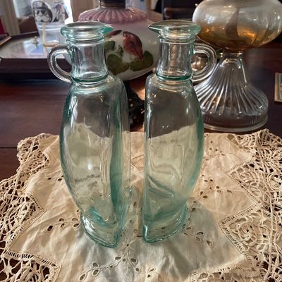 Sale Photo Thumbnail #40: Pair of vintage light green glass oil and vinegar cruets with small side handles, displayed on a lace doily.