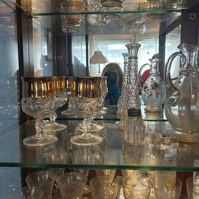 Sale Photo Thumbnail #56: Collection of vintage glass stemware, decanters, and crystal pieces displayed on glass cabinet shelves, including gold-rimmed goblets and etched wine glasses.