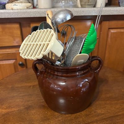 Sale Photo Thumbnail #17: Brown glazed stoneware crock filled with assorted vintage and modern kitchen utensils, including a ladle, hand grater, whisk, and plastic spatulas.