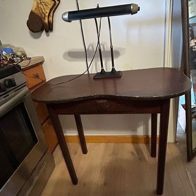 Sale Photo Thumbnail #121: Vintage dark wood kidney-shaped writing desk featuring a center drawer, paired with a black metal double-arm piano lamp.