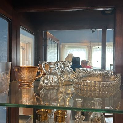 Sale Photo Thumbnail #57: Assorted vintage glassware displayed in a mirrored cabinet, including an amber depression glass cup, clear glass cruets, cut crystal trays, and gold-rimmed goblets.