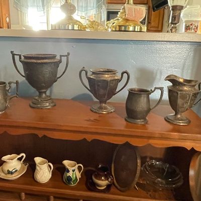 Sale Photo Thumbnail #3: Collection of tarnish-patinated pewter and silver-plate vessels displayed alongside small ceramic creamers and glassware on a wooden shelf.