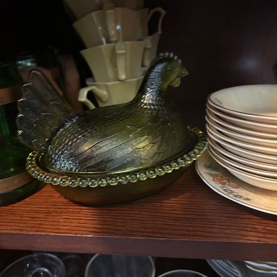 Sale Photo Thumbnail #54: Vintage olive green glass hen-on-nest covered dish displayed inside a dark wooden cabinet, flanked by stacks of dinnerware.