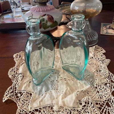 Sale Photo Thumbnail #41: Pair of vintage light blue glass bottles with small side handles, sitting on a crocheted lace doily alongside oil lamps.