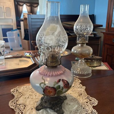 Sale Photo Thumbnail #43: Antique hand-painted floral oil lamp with an etched glass chimney, resting on a lace doily alongside a clear glass kerosene lamp.