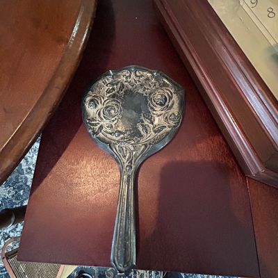 Sale Photo Thumbnail #34: Vintage ornate silver-plated hand mirror featuring embossed rose floral details, resting on a dark wood surface.