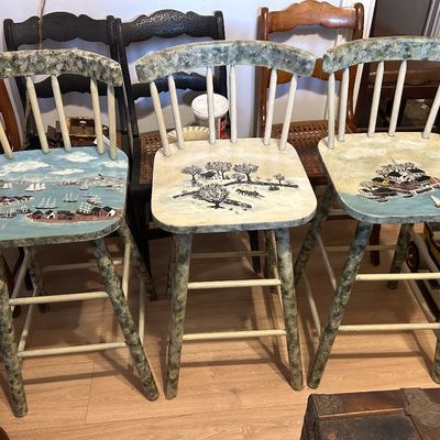 Sale Photo Thumbnail #107: Trio of wooden bar stools with spindle backs, featuring hand-painted folk art scenes of coastal and winter landscapes on the seats.