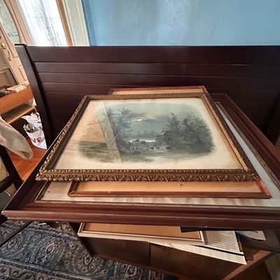 Sale Photo Thumbnail #37: Stack of framed artwork including a antique landscape print in an ornate gilt wood frame resting on a dark wood headboard.