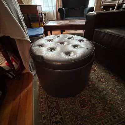 Sale Photo Thumbnail #117: Round, dark brown faux-leather storage ottoman with a button-tufted top cushion.