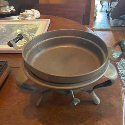 Sale Photo Thumbnail #65: Vintage metal chafing dish set on a four-legged warming stand, featuring dark side handles and an adjustable burner knob beneath.