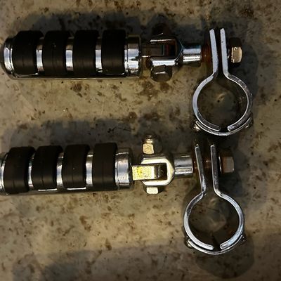 Sale Photo Thumbnail #144: Pair of chrome motorcycle highway foot pegs with ribbed rubber grips and frame clamps, showing light surface wear and minor oxidation.