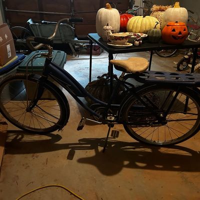 Sale Photo Thumbnail #123: Vintage dark blue cruiser bicycle with rear rack, parked in front of a table holding seasonal pumpkins and decor.