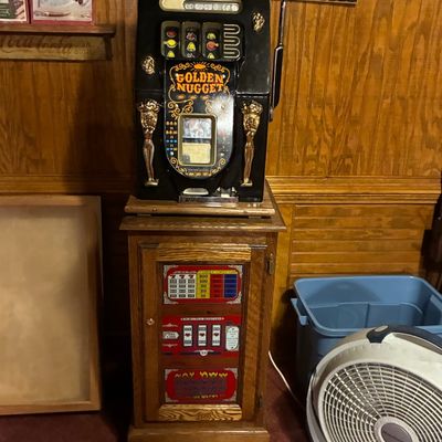 Sale Photo Thumbnail #185: Vintage Golden Nugget 25-cent slot machine on a matching wooden cabinet stand, featuring ornate gold-tone figures and classic three-reel display.