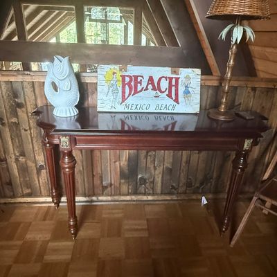 Sale Photo Thumbnail #240: Polished mahogany console table featuring turned legs and gold detailing, topped with a white fish vase, painted wood 'Mexico Beach' sign, and a tropical palm tree table lamp.