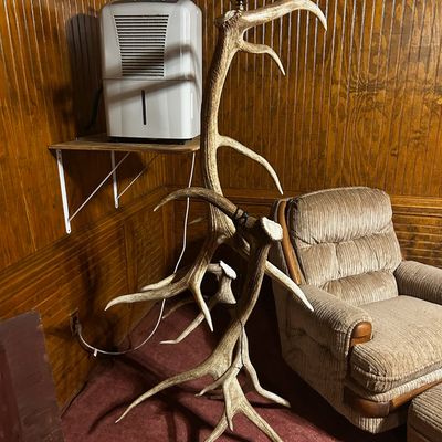 Sale Photo Thumbnail #132: Rustic floor lamp constructed from interlocking deer antlers, lacking a lampshade, shown standing next to a plush recliner in a wood-paneled room.