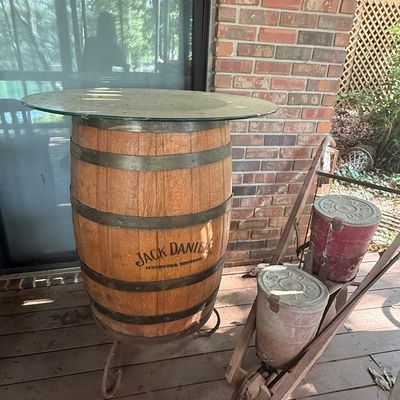 Sale Photo Thumbnail #225: Jack Daniel's wooden whiskey barrel table with a round glass top, alongside a vintage rustic wooden planter or cart.