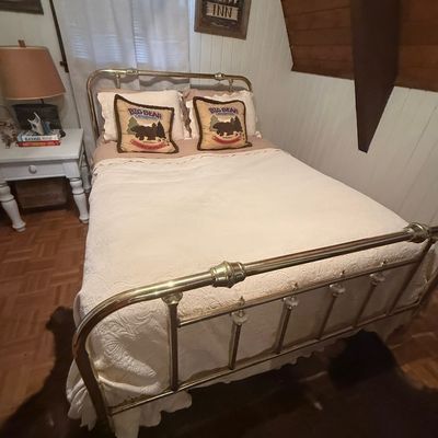 Sale Photo Thumbnail #223: Vintage brass full-size bed frame featuring decorative ceramic spindles on the headboard and footboard. Includes a white quilted bedspread and rustic cabin-themed decorative pillows.