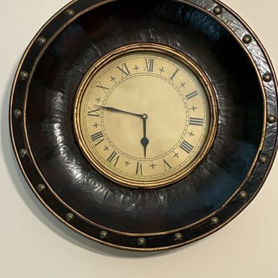 Sale Photo Thumbnail #211: Round wall clock with a dark faux-leather frame accented by brass stud detailing and a beige face with Roman numerals.