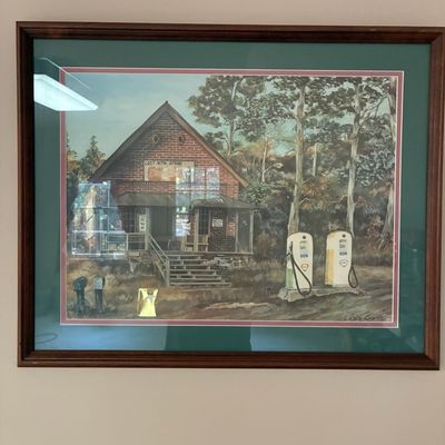 Sale Photo Thumbnail #192: Framed and matted art print depicting a rustic general store with vintage gas pumps, signed by artist Leslie Grant.