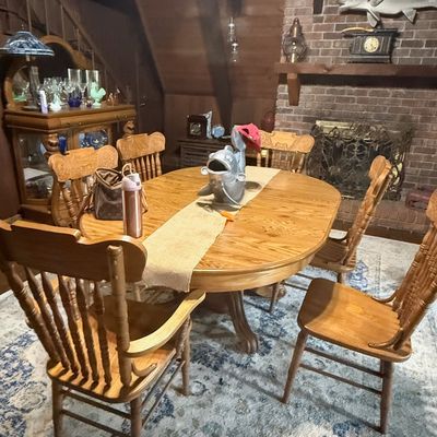 Sale Photo Thumbnail #243: Traditional oak pedestal dining table with six matching pressed-back spindle chairs set on a patterned area rug in a rustic dining room.