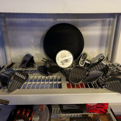 Sale Photo Thumbnail #19: Assortment of motorcycle side mirrors, chrome hardware, and a Harley-Davidson branded round cover arranged on a white plastic storage shelf.