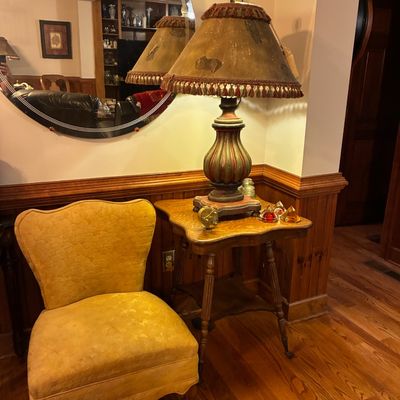 Sale Photo Thumbnail #180: Vintage mustard yellow upholstered accent chair paired with a wooden side table featuring a painted urn-style table lamp with a fringed shade.