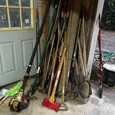 Sale Photo Thumbnail #39: Assortment of long-handled yard tools including a red-headed axe, shovels, hoes, and a gas-powered pole saw leaning against a garage wall.