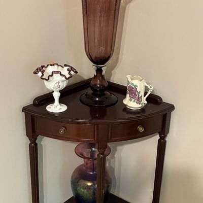 Sale Photo Thumbnail #208: Traditional mahogany-finish corner table displaying a tall amethyst glass floor vase, Fenton hand-painted milk glass compote, ceramic grape-pattern pitcher, and an iridescent carnival glass vase.